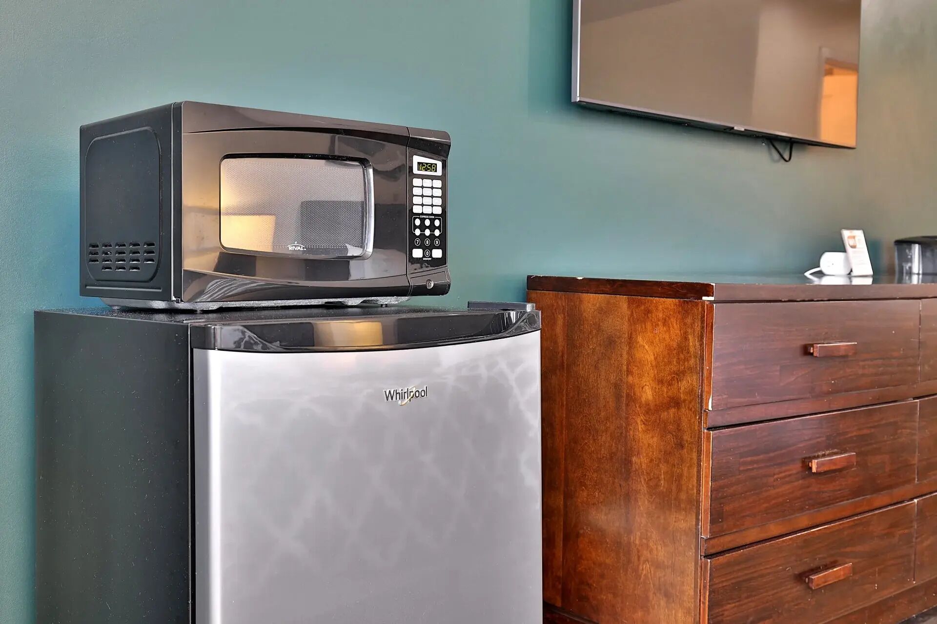 Un refrigerador compacto con un microondas encima, un aparador de madera con cajones y un televisor montado en la pared.