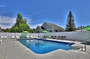 Una piscina con sillones de descanso y sombrillas, rodeada por una valla blanca y altos árboles.