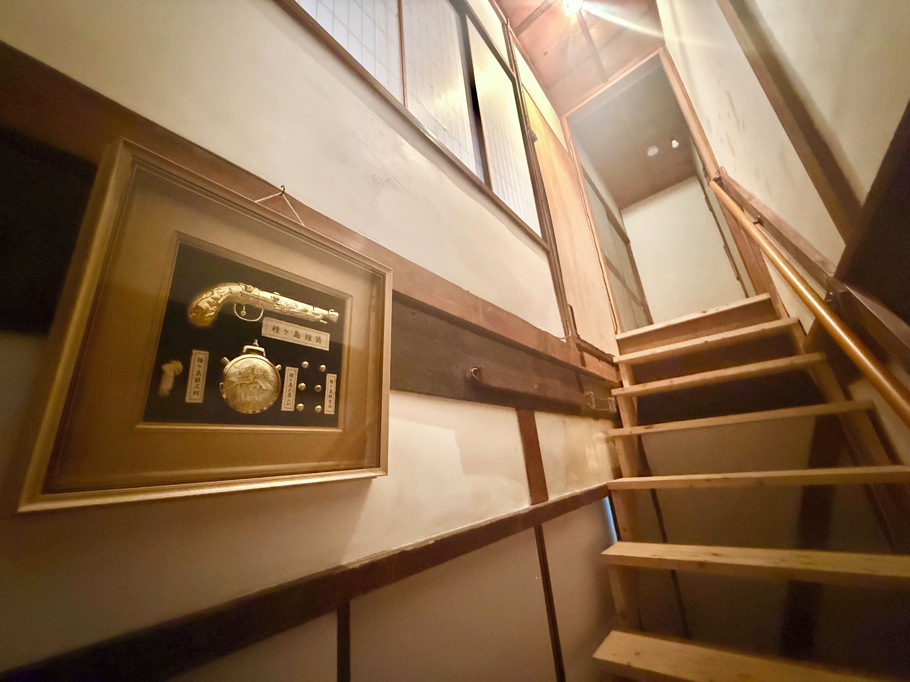 A framed display with a clock and other items on a wall, a wooden staircase with a handrail, and a window above.