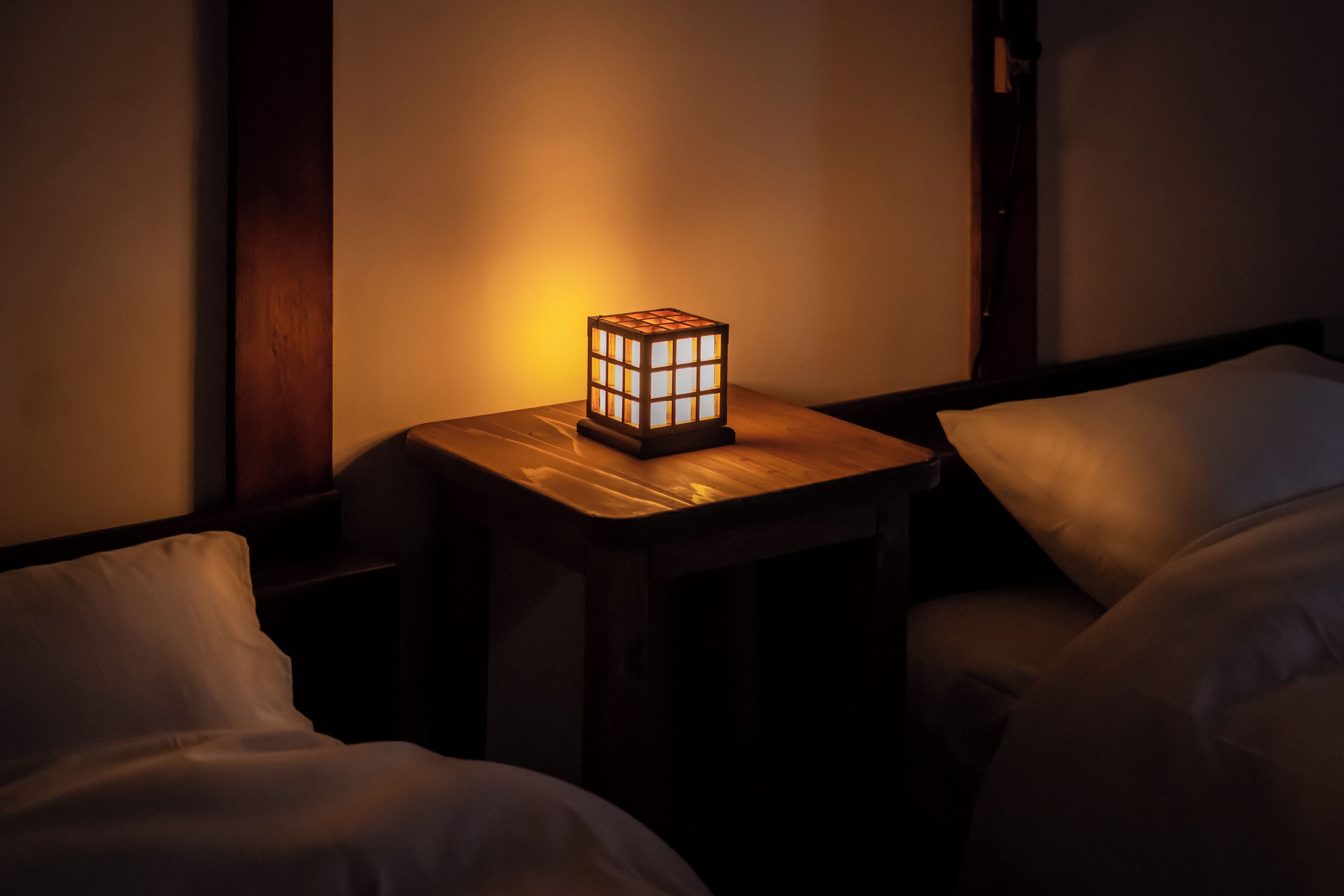 A room with a bedside table featuring a cube-shaped lamp, a wooden nightstand, and a bed with white bedding.