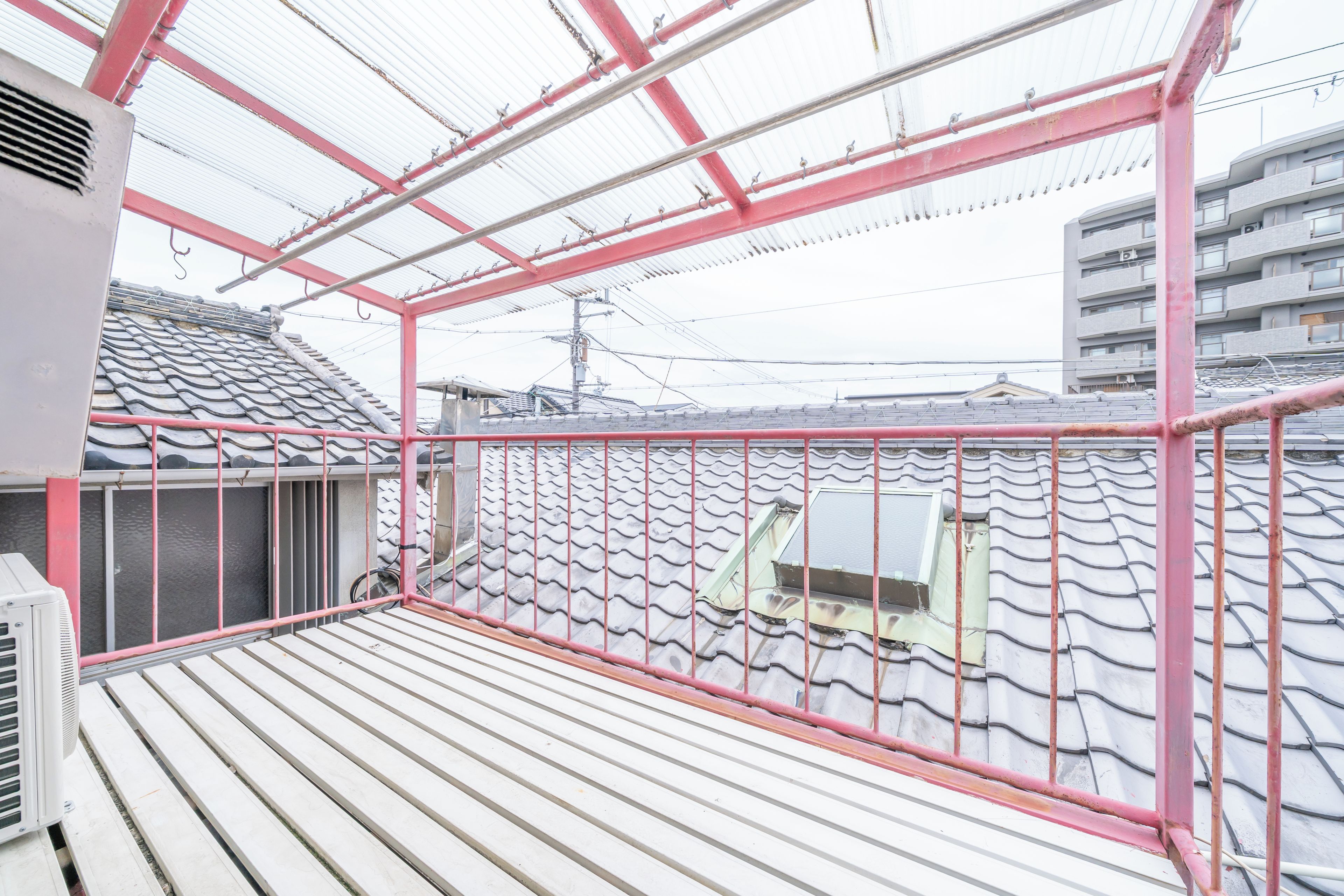 A balcony with a tiled roof, a red railing, and a view of a residential building.