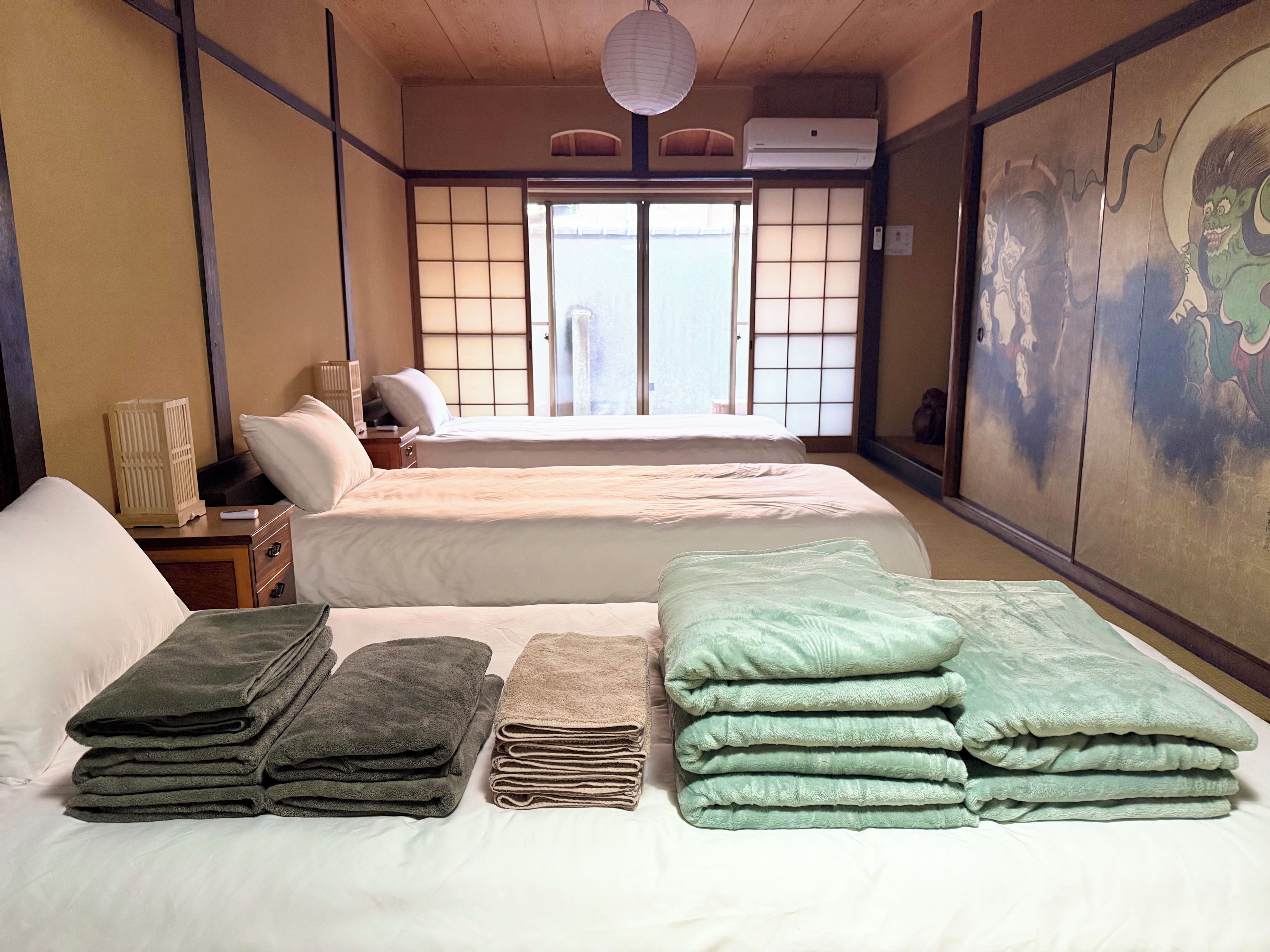A traditional Japanese-style room with tatami flooring, a shoji screen door, and a futon bed. There are folded towels in green and beige, and a small nightstand with a lamp.