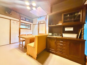 Una habitación con una mesa de madera, sillas, un armario con cajones, una ventana y una lámpara en el techo.