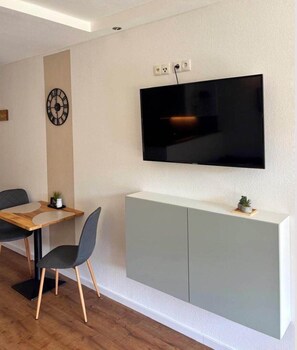 Un salon moderne avec un téléviseur à écran plat fixé au mur, une table à manger avec des chaises et un meuble gris placé sous le téléviseur.