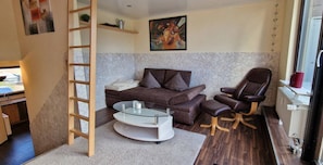 A compact living space with a sofa, a glass coffee table, a chair, a ladder, a radiator, and a painting on the wall.