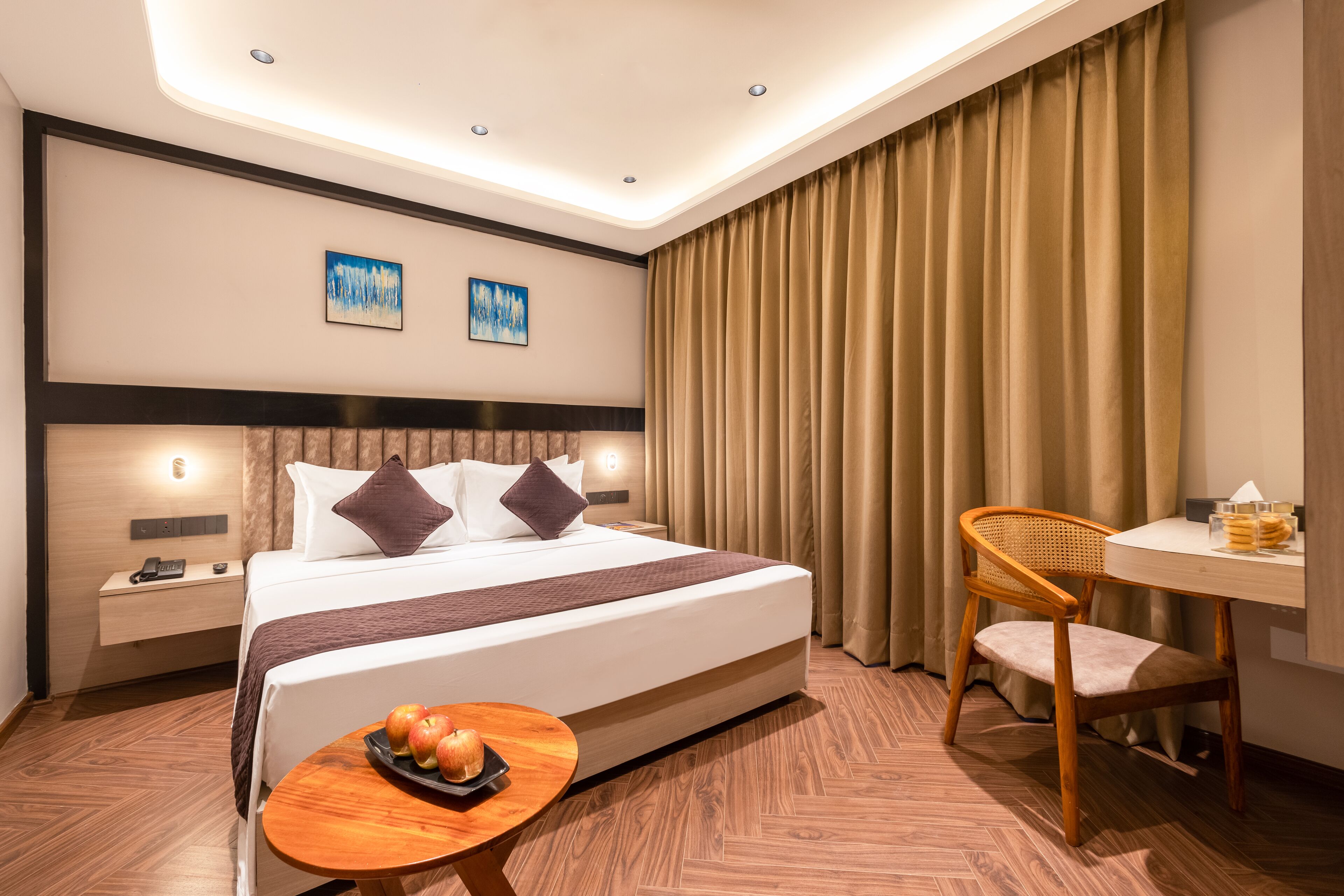 Una habitación de hotel con una cama grande, una mesita de noche de madera, una pequeña mesa con manzanas, una silla y un escritorio con una lámpara.