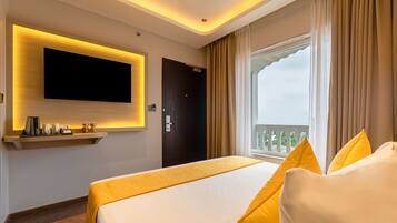 Hotelkamer met groot bed, wandtelevisie, raam met gordijnen en balkonuitzicht.