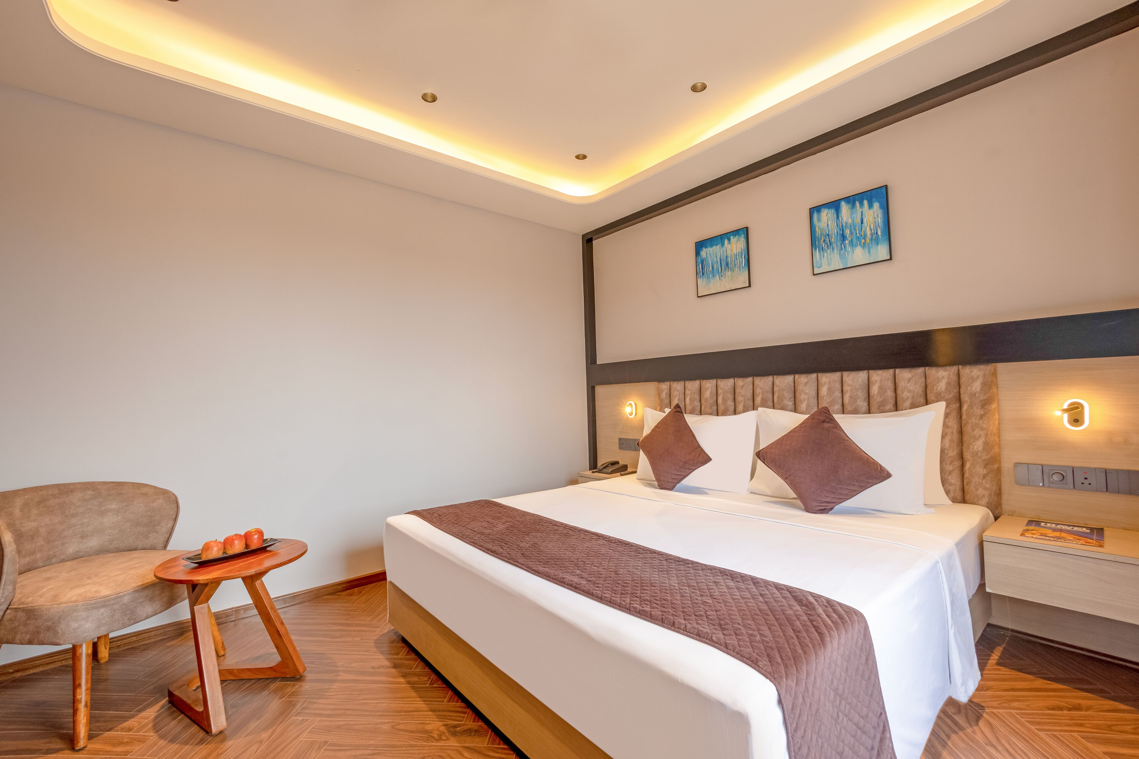 A modern hotel room with a large bed, a wooden side table, a chair, and a small round table with fruit.