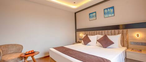 A modern hotel room with a large bed, a wooden side table, a chair, and a small round table with fruit.