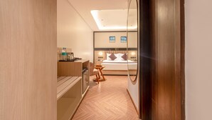 Une chambre d’hôtel moderne avec un plancher en bois, un lit avec une literie blanche et deux oreillers, une table de chevet avec une lampe, une petite chaise en bois et un bureau avec une chaise. Un grand miroir est accroché au mur et un luminaire est fixé au plafond.