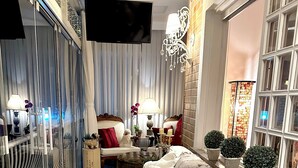Una habitación acogedora con sofá, una mesita y televisión. Hay plantas y una ventana con cortinas.