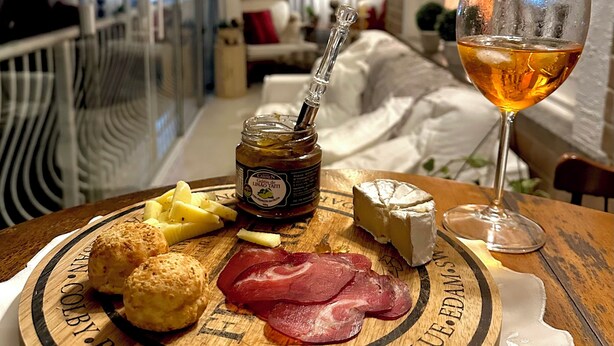 Una tabla de madera con queso, jamón y galletas sobre una mesa, junto a una copa de vino y una botella de mermelada.
