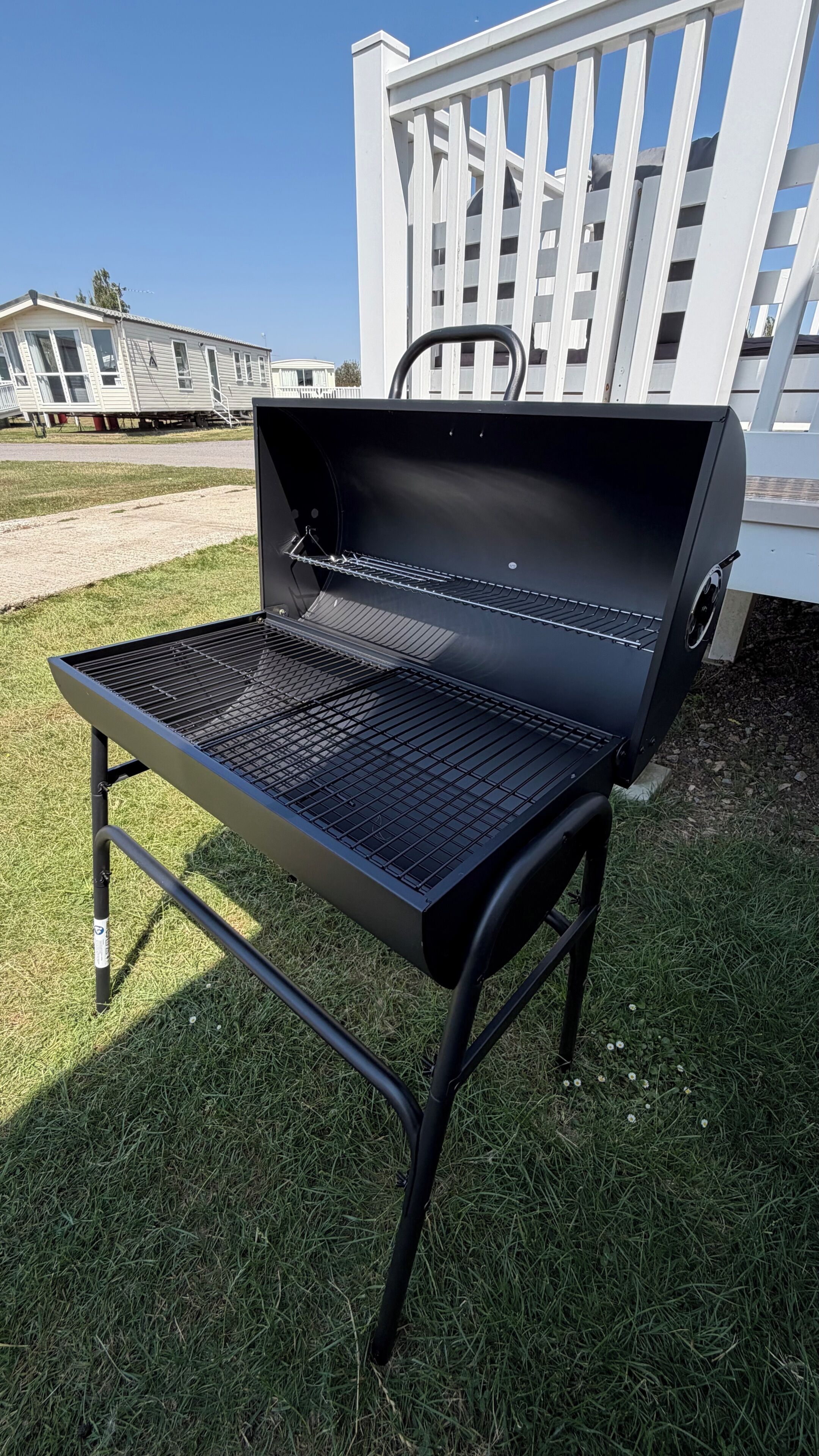 En sort grill med metalrist, placeret på en græsplæne med hvidt hegn og et hus i baggrunden.