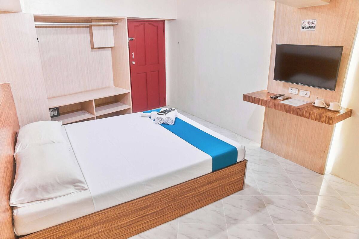 Ein Hotelzimmer mit Bett, Fernseher, Schreibtisch mit Tasse und Fernbedienung, roter Tür und einem Rauchverbot-Schild.