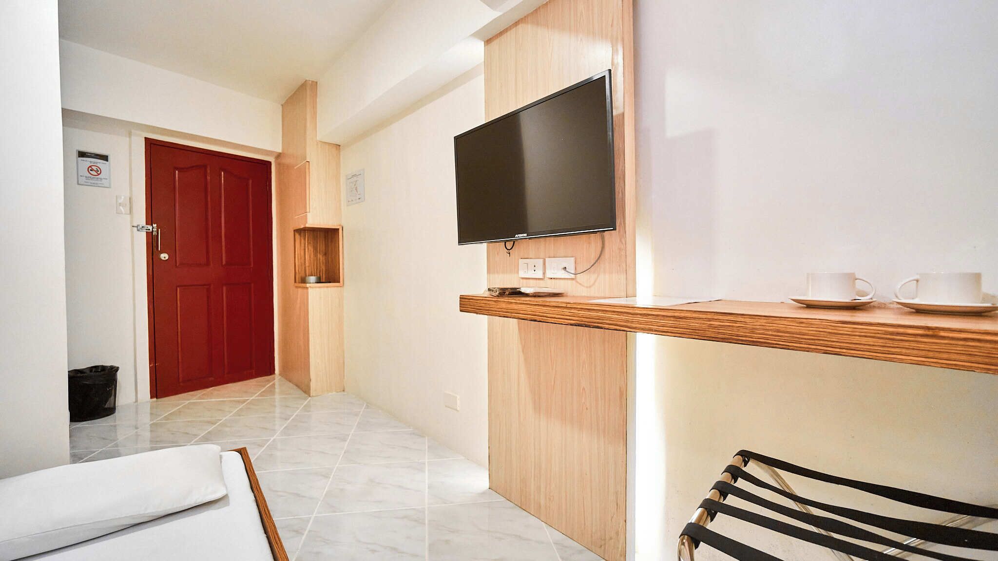 A hotel room with a TV mounted on the wall, a wooden shelf with cups, a red door, and a tiled floor.