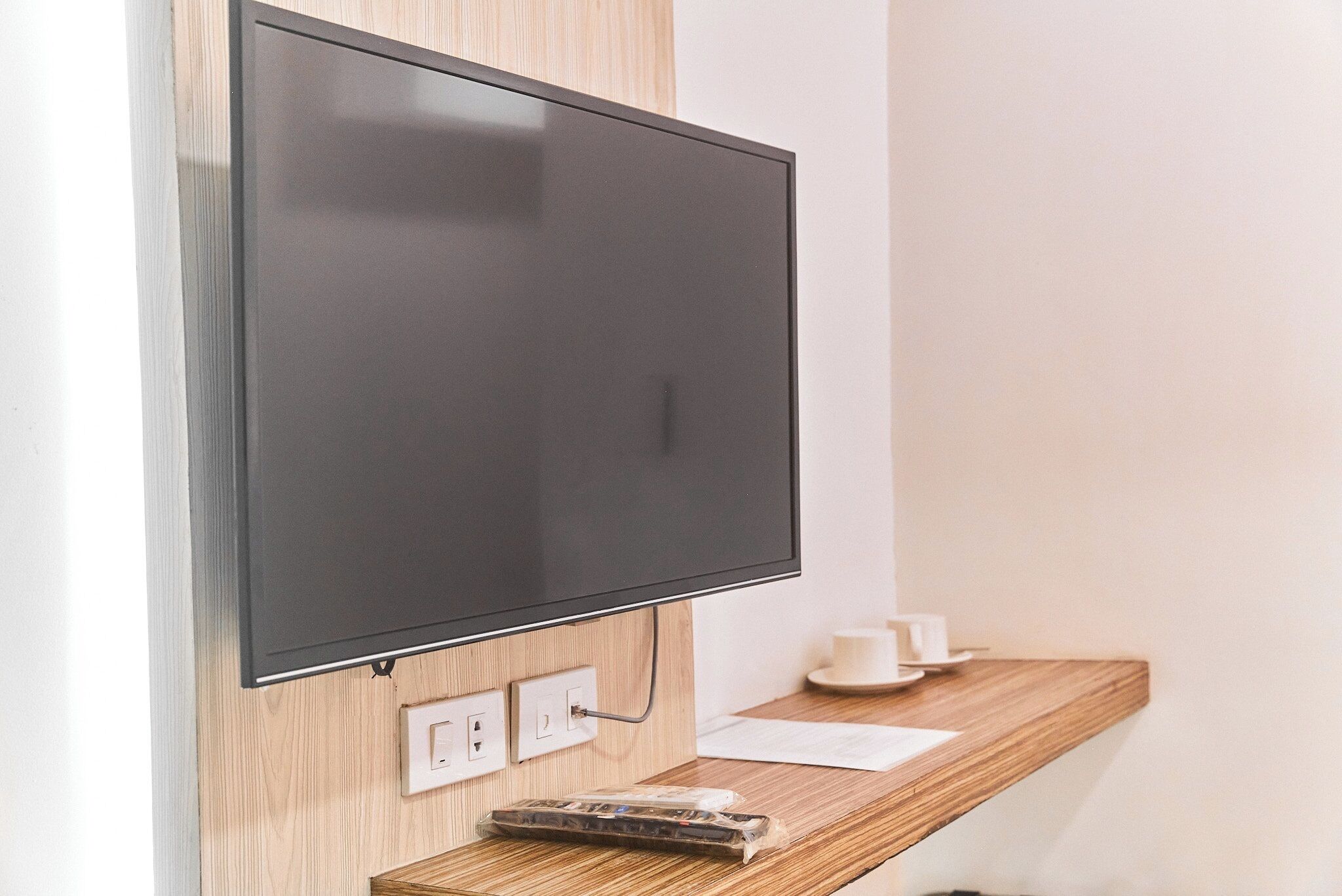 Ein Flachbildfernseher an einer Holzwand, eine Steckdose und ein kleiner Tisch mit einer Tasse und einer Fernbedienung.