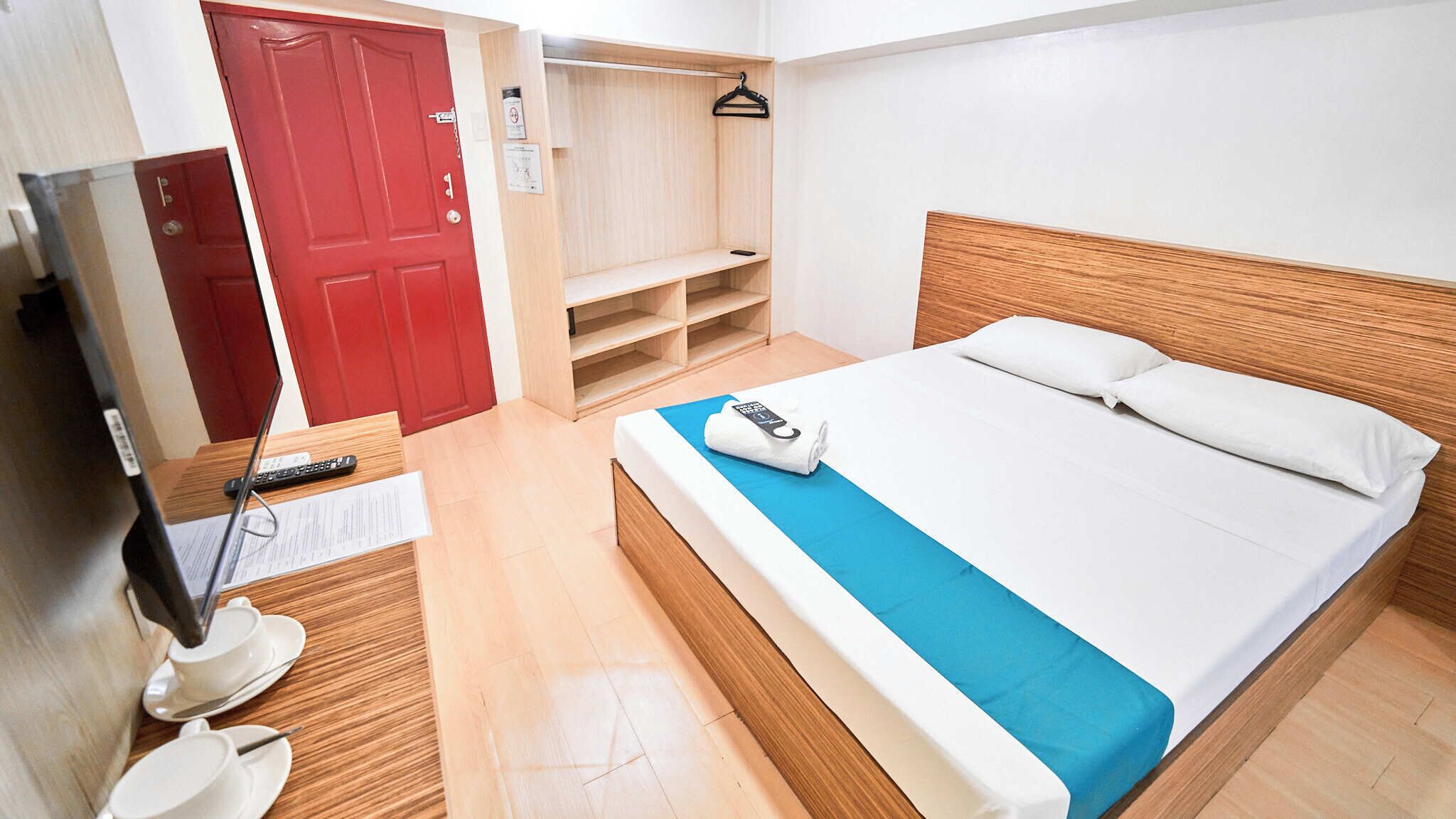Habitación de hotel con una cama, un televisor, una puerta roja, un armario de madera y una colcha azul.