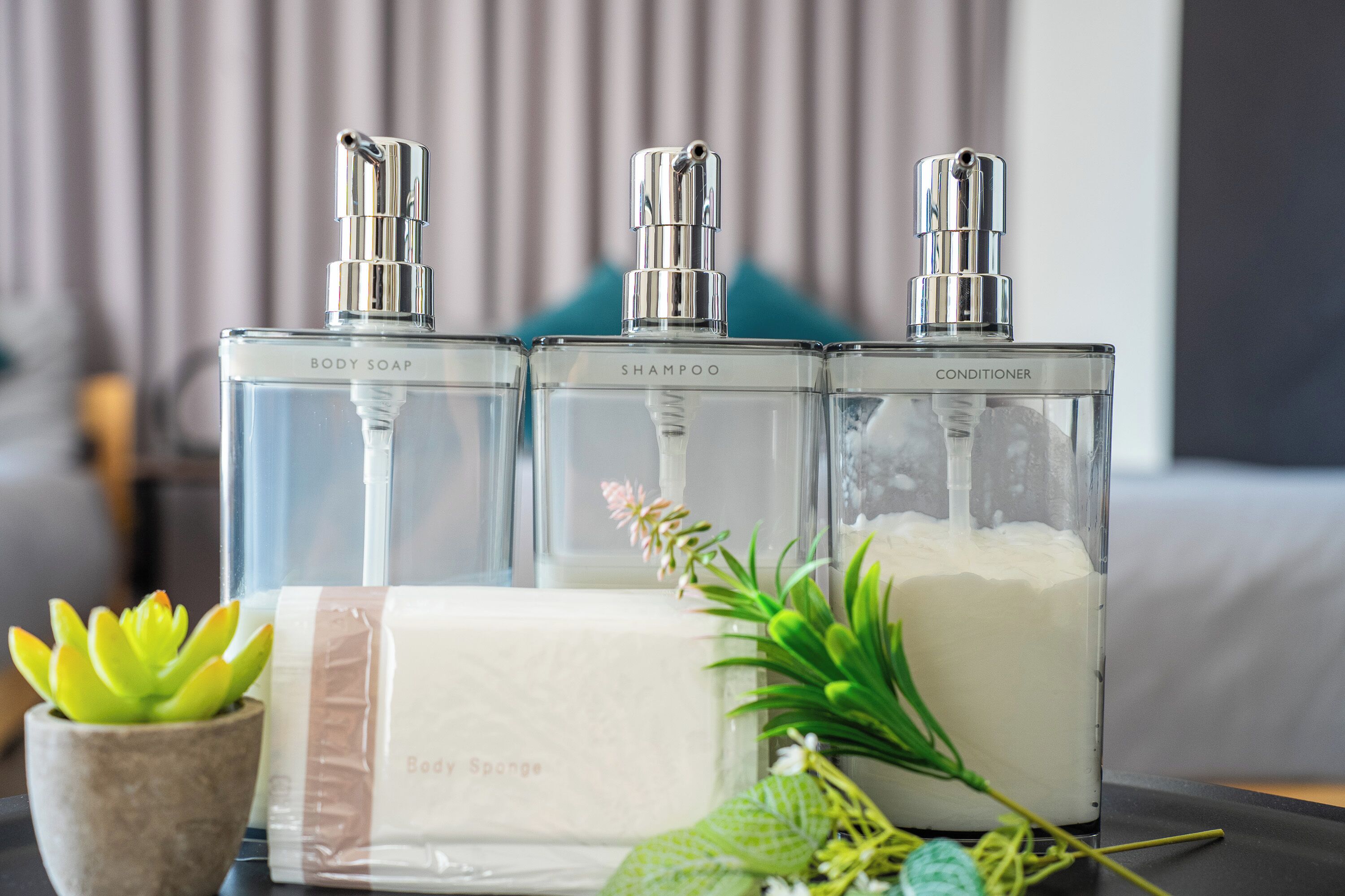 Tres botellas de vidrio transparente etiquetadas con jabón corporal, champú y acondicionador sobre una mesa, junto con una esponja de baño y plantas decorativas.