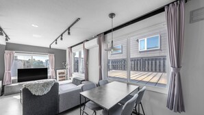 Una sala de estar moderna con sofá, mesa de comedor y sillas, ventanas amplias con cortinas y un televisor de pantalla plana.