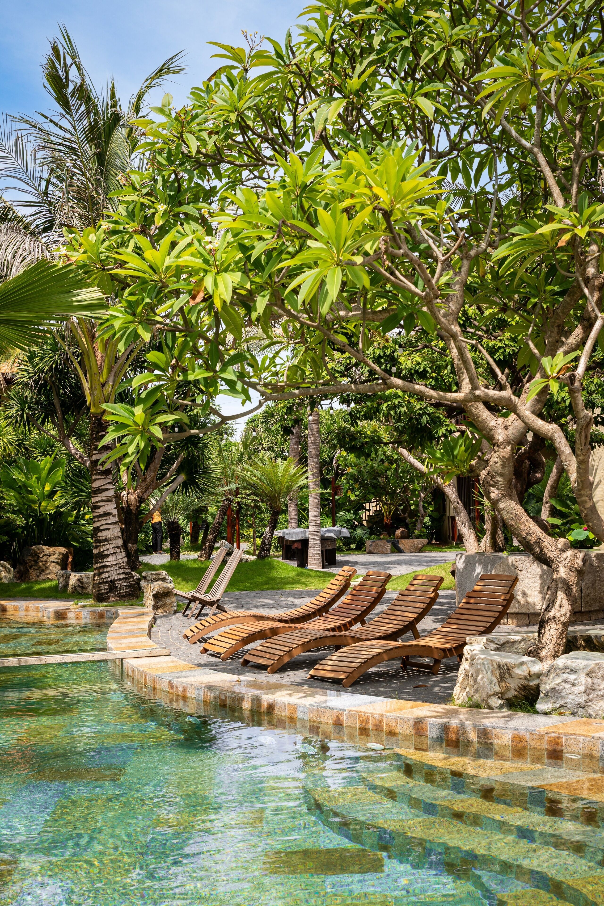 A pool area with clear water, surrounded by lush greenery and wooden lounge chairs.