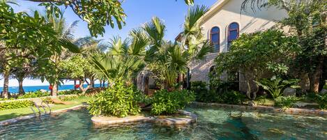 Un espace piscine avec une eau cristalline, entouré de plantes tropicales et bordé d’un bâtiment doté de fenêtres en arc de cercle.