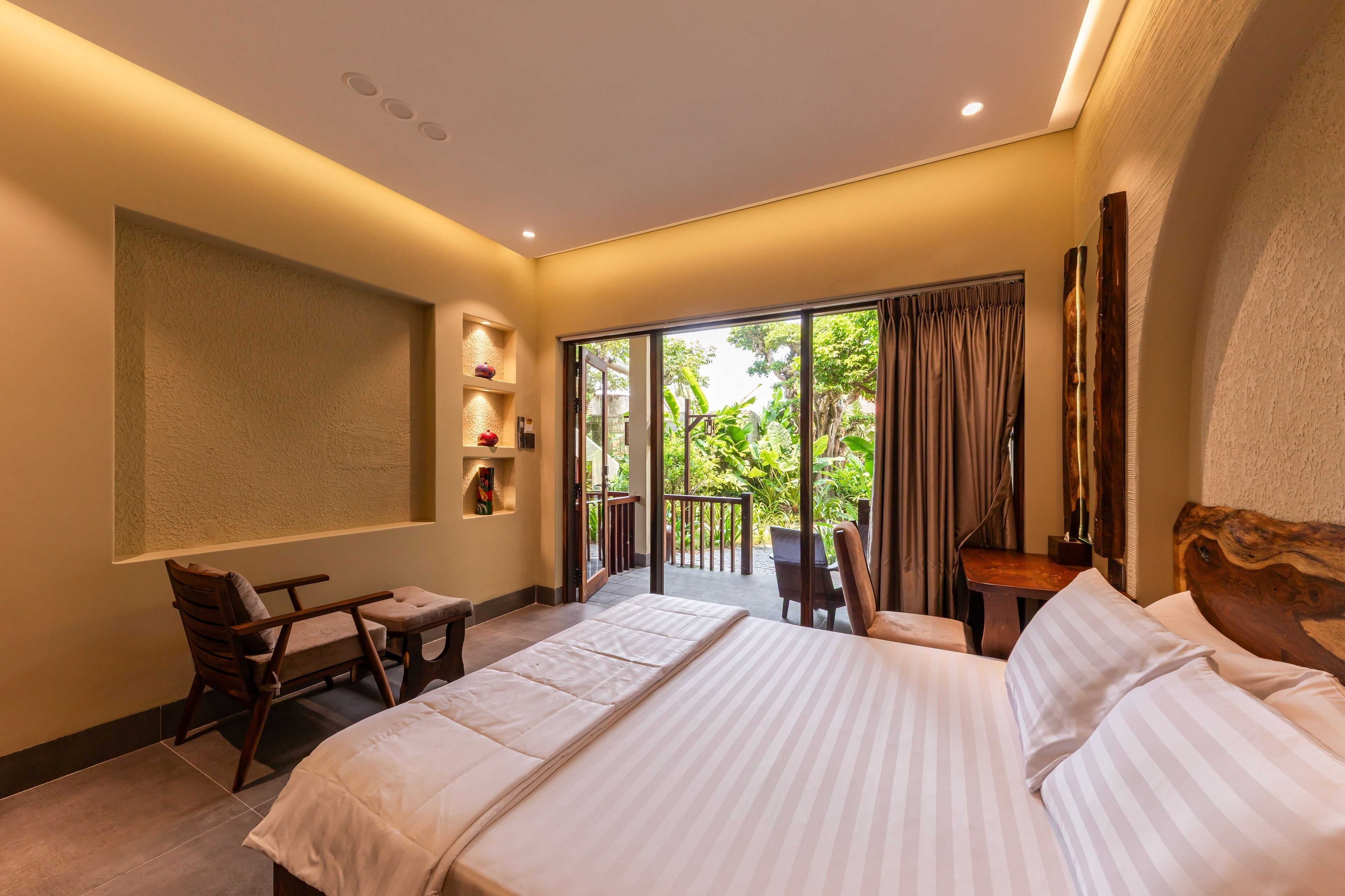 A hotel room with a large bed, a small desk, a chair, and a balcony with a view of greenery.