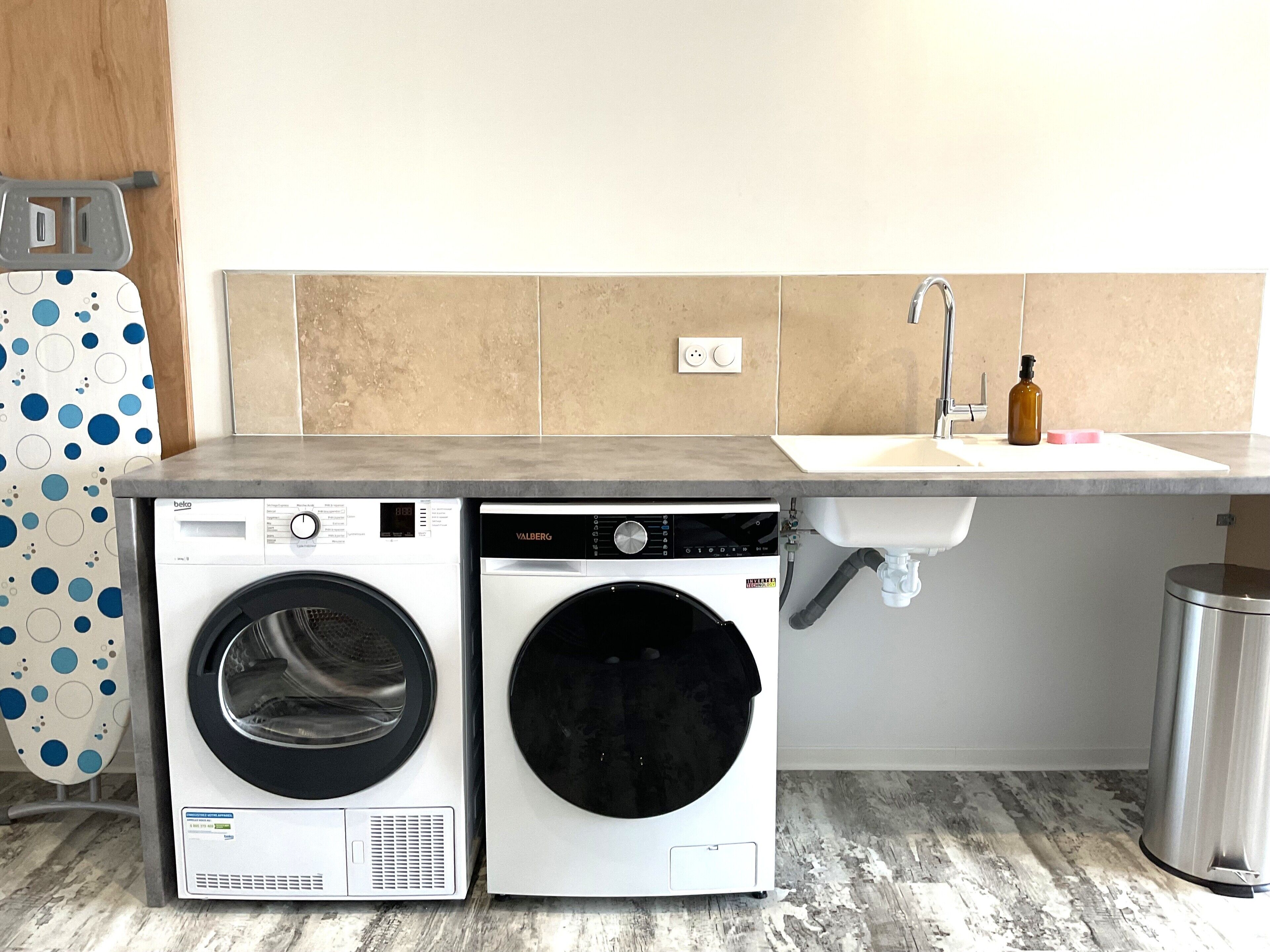 A compact laundry area with two front-loading washing machines, a sink, and a trash can.