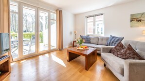 A spacious living room with a wooden coffee table, a grey sofa, and a television on a wooden stand. Large windows provide ample natural light and a view of the outdoors.