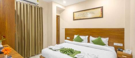 A hotel room with a wooden headboard, a bed with white linens, green pillows, a framed quote on the wall, an air conditioning unit, and a wooden nightstand with a phone.