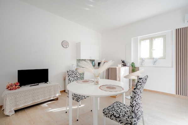 A modern living room with a flat-screen TV, a white round dining table with black and white patterned chairs, a white cabinet, and a window with vertical blinds.