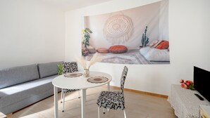 Ein Wohnzimmer mit grauem Sofa, einem runden weißen Tisch, gemusterten Stühlen, einer Wanddekoration aus Makramee und einem Fernseher auf einem Beistelltisch.