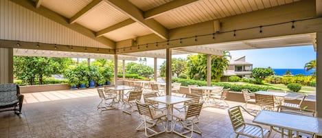 Un área de patio techada al aire libre con varias mesas y sillas, rodeada de follaje y vistas al océano.