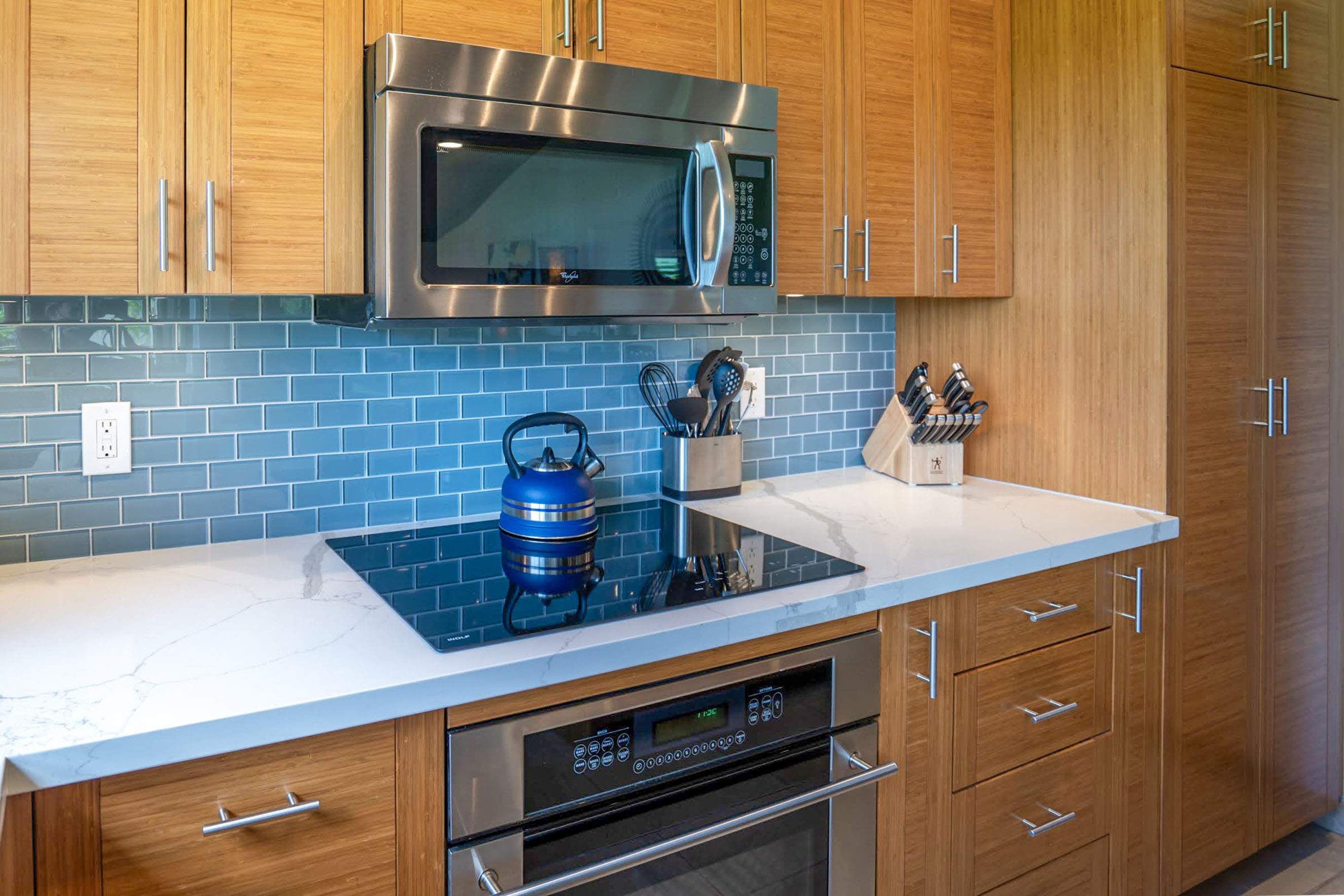 A modern kitchen with wooden cabinets, a stainless steel microwave, a blue kettle, and a built-in oven.