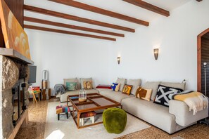 A living room with a fireplace, wooden beams, a sofa, a coffee table, and a green ottoman.