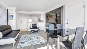 A modern living room with a sofa, a glass dining table, and chairs. There is a kitchen in the background with white cabinets and a marble backsplash. - Azure Tides Oceanfront (Daytona Beach)