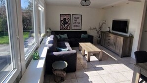 A spacious living room with a large sofa, a coffee table, and a TV. There are two framed pictures on the wall and a window with blinds.