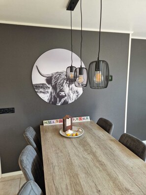 Un espace repas comprenant une table en bois, des chaises à revêtement gris, une décoration murale ronde représentant un chien et des lampes suspendues.