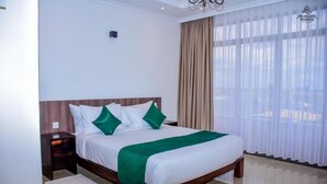 A hotel room with a large bed, a wooden headboard, and a view through sheer curtains. - Highland Rhinor (Eldoret)