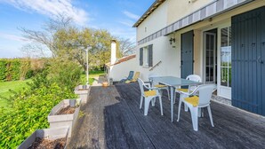 A patio with a table and chairs, a white building with a dark door, and a garden with bushes. - Renovated Holiday Home 'Le 36' with Private Terrace, Garden, and Wi-Fi (Jonzac)