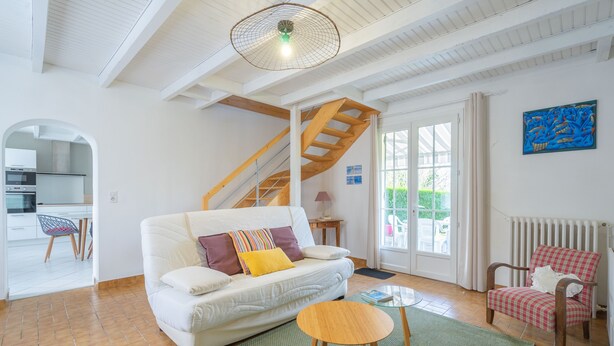 A living room with a white sofa, a wooden coffee table, a red and white checkered chair, a staircase, and a kitchen visible through an archway. - Renovated Holiday Home 'Le 36' with Private Terrace, Garden, and Wi-Fi (Jonzac)