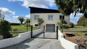Une maison à deux étages, avec un toit noir, des murs blancs et une allée de gravier menant à une entrée fermée par un portail.