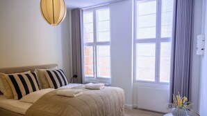 Un letto rifatto con cuscini a righe, un comodino con un vaso di fiori e ampie finestre con tende.