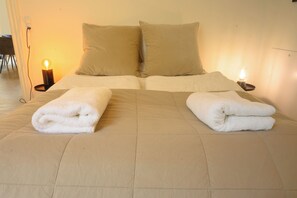 A neatly made bed with two pillows and two folded towels, a bedside lamp, and a power outlet on the wall.
