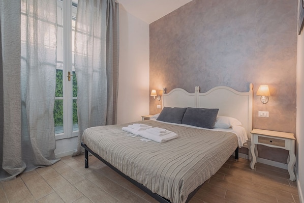 A bedroom with a bed, two bedside tables, a nightstand, a window with curtains, and a wall-mounted lamp.