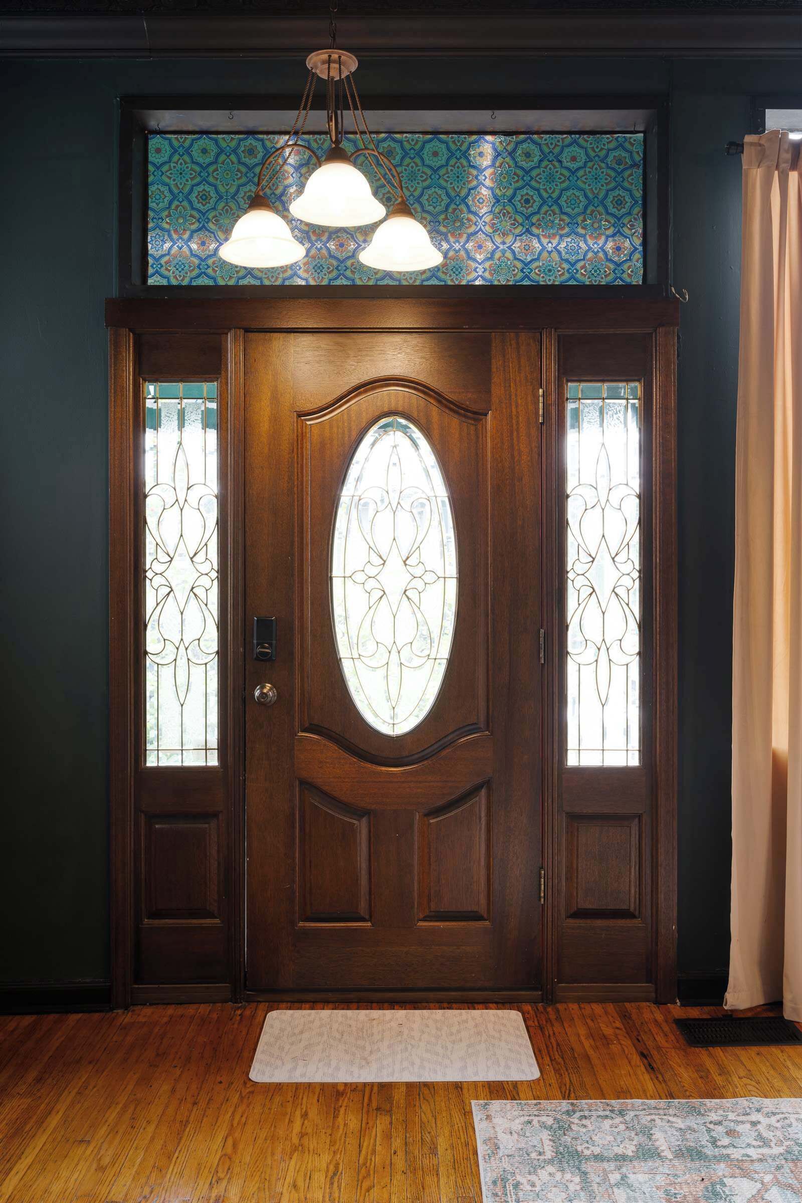Une porte d’entrée en bois, ornée de panneaux de vitraux et surmontée d’un luminaire décoratif au plafond.