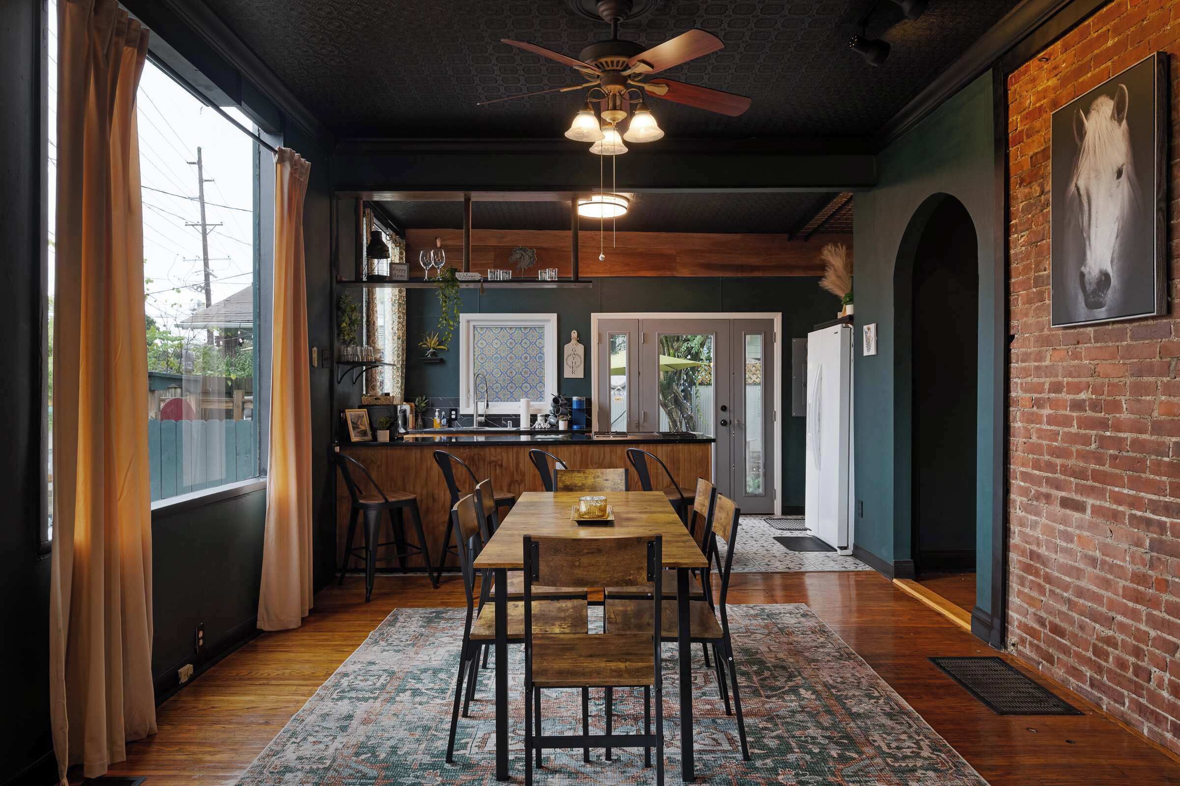 Un'area pranzo con un tavolo e sedie in legno, una ventola a soffitto e vista sulla cucina e sala da pranzo attraverso una porta aperta.