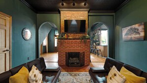 A living room with a brick fireplace, two leather sofas with yellow cushions, a large mirror, and a painting of a tree. - Retreat on Bardstown Road | Yard • Great Location (Louisville)