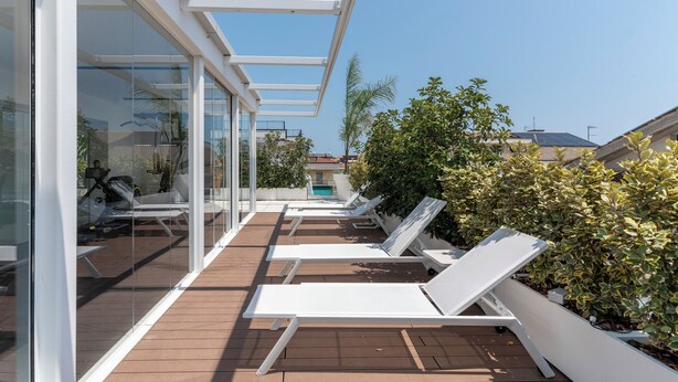 Une terrasse sur le toit, dotée de chaises longues blanches, d’un espace clos par des parois de verre et offrant une vue sur le quartier environnant.