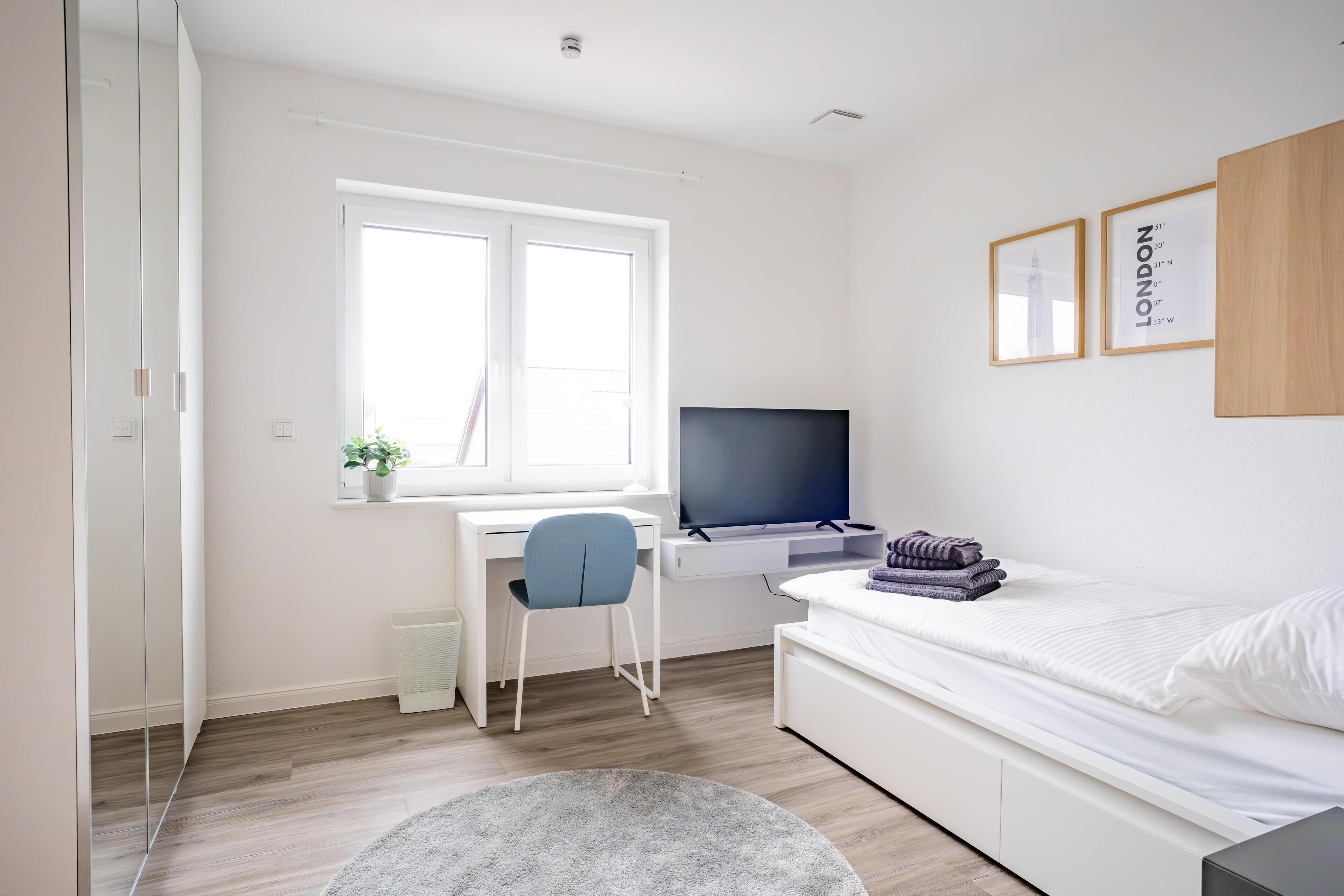A modern bedroom with a bed, a desk, a chair, a TV, and a window.
