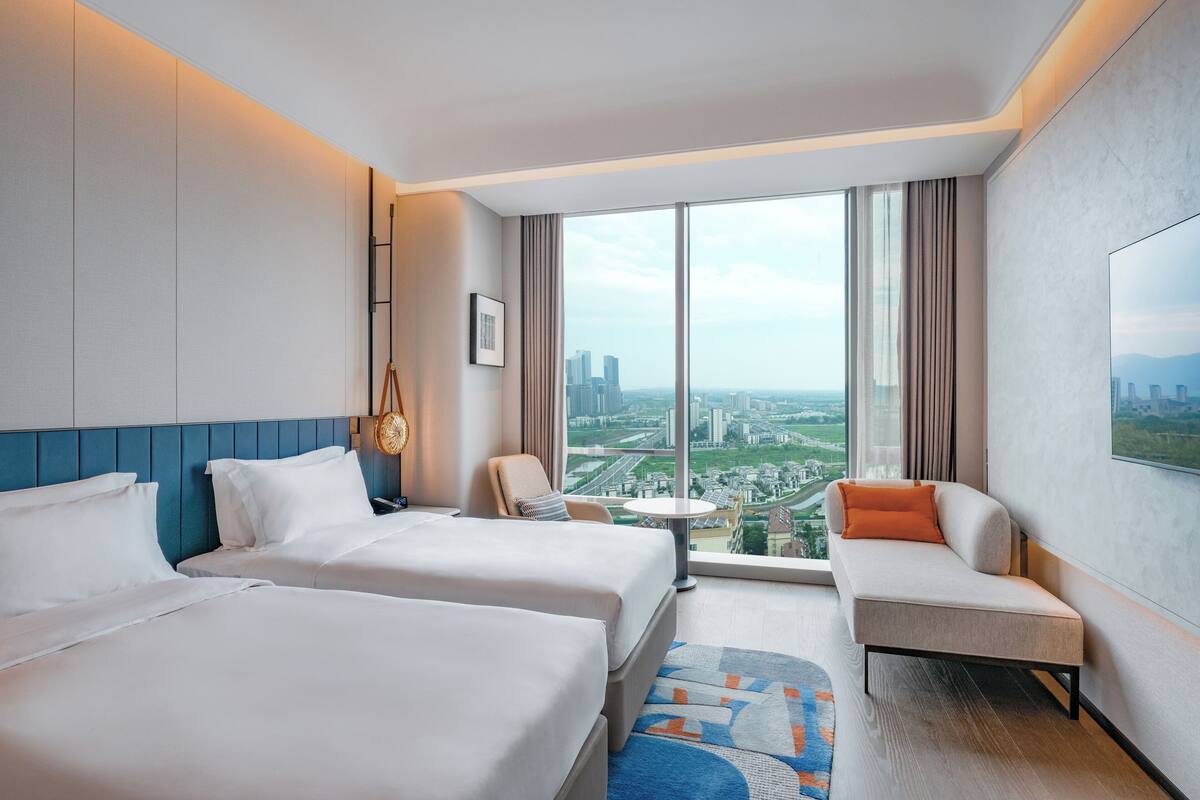 Habitación de hotel con dos camas, un sofá, una mesita pequeña y un ventanal amplio con vistas a la ciudad.